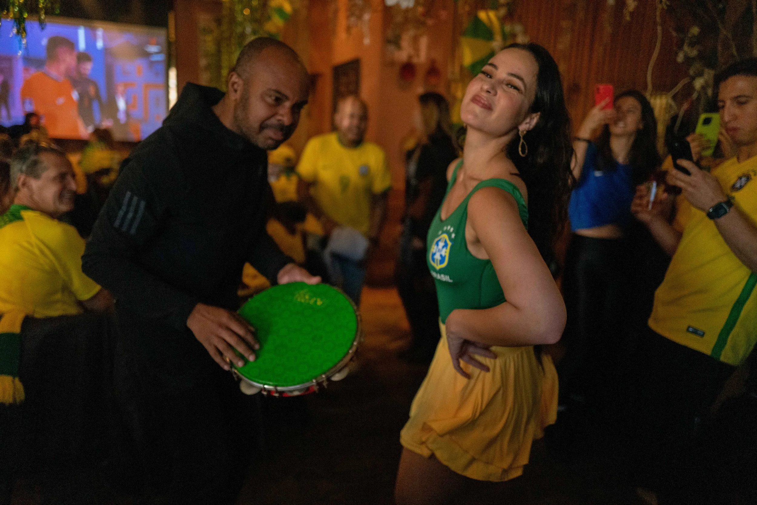 A woman in a green dress with the Brazil national team logo dancing at a lively party with people wearing yellow shirts, holding a tambourine, and taking photos in a warmly lit room.
