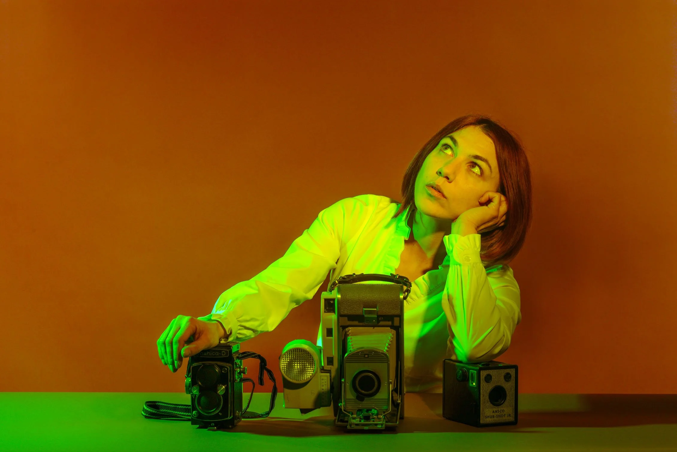 A woman with red hair and a white shirt is sitting at a table with vintage cameras and a flash in front of her. She is resting her head on her left hand and looking upward with a thoughtful expression. The background is a gradient of warm orange and brown. The lighting has a greenish tint.