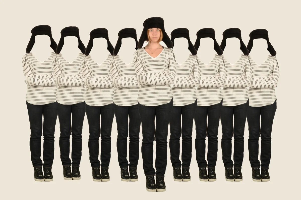 A woman wearing a black winter hat standing in front of a line of eight faceless women dressed identically in striped shirts and black pants, all with their arms crossed.