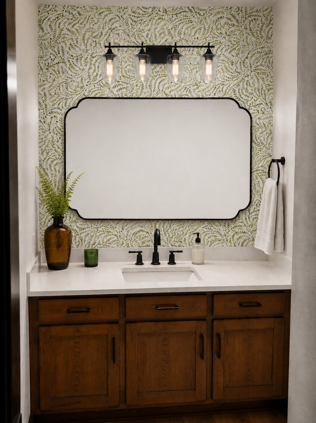 Bathroom vanity with a large mirror framed with black, a wall-mounted light fixture with four bulbs, a brown vase with green leaves, a small green candle holder, a soap dispenser, and a black towel ring with a white towel. The vanity has a white coun