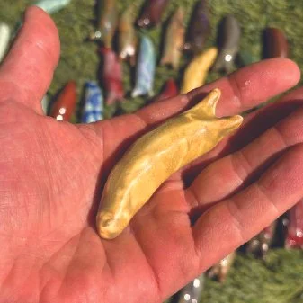 A hand holding a small yellow clay slug with a blurred background of various other colorful slugs on grass.