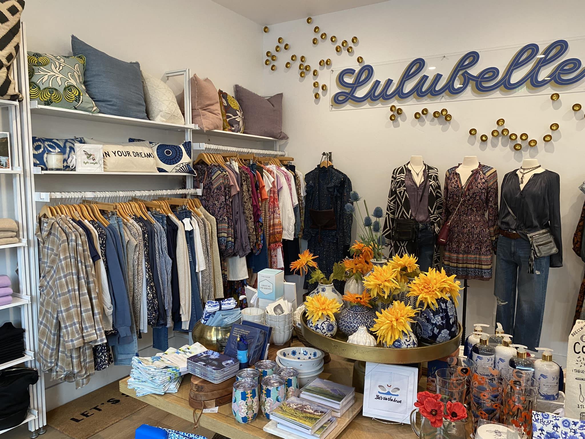 Interior of a retail store displaying clothing, pillows, home decor, and accessories, with a sign that says 'Lulu belle' on the wall.