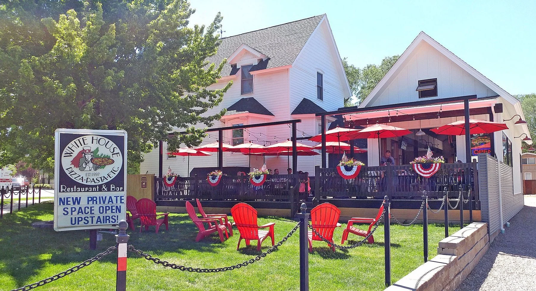 Exterior view of White House Pizza restaurant with outdoor seating and red umbrellas, decorated with patriotic bunting, on a sunny day. There are chairs in the grass area and a sign indicating new private space upstairs.