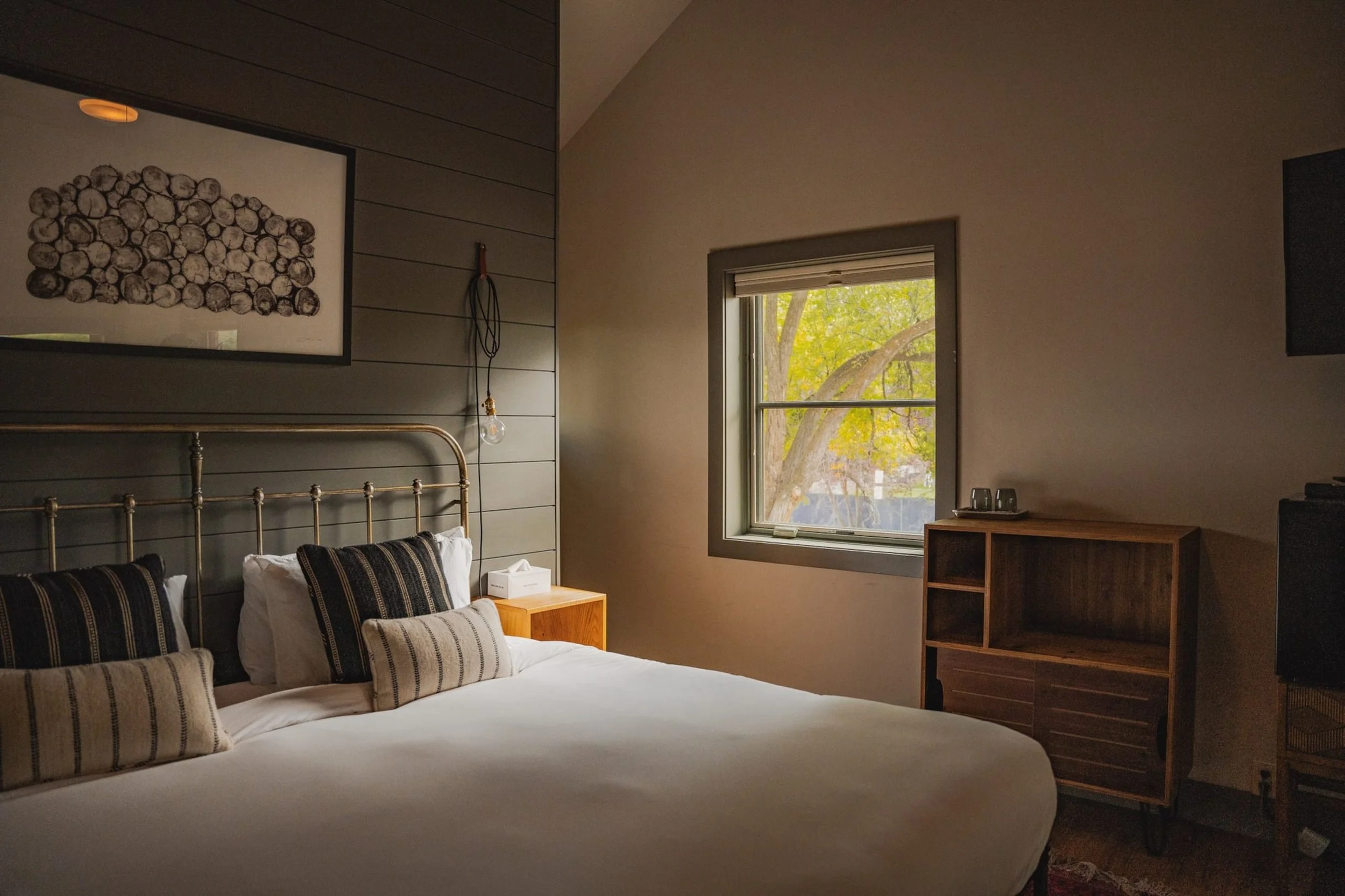 A cozy bedroom with a large bed that has striped pillows, a wooden nightstand, a window showing trees outside, and a framed art piece of stacked logs on the wall.