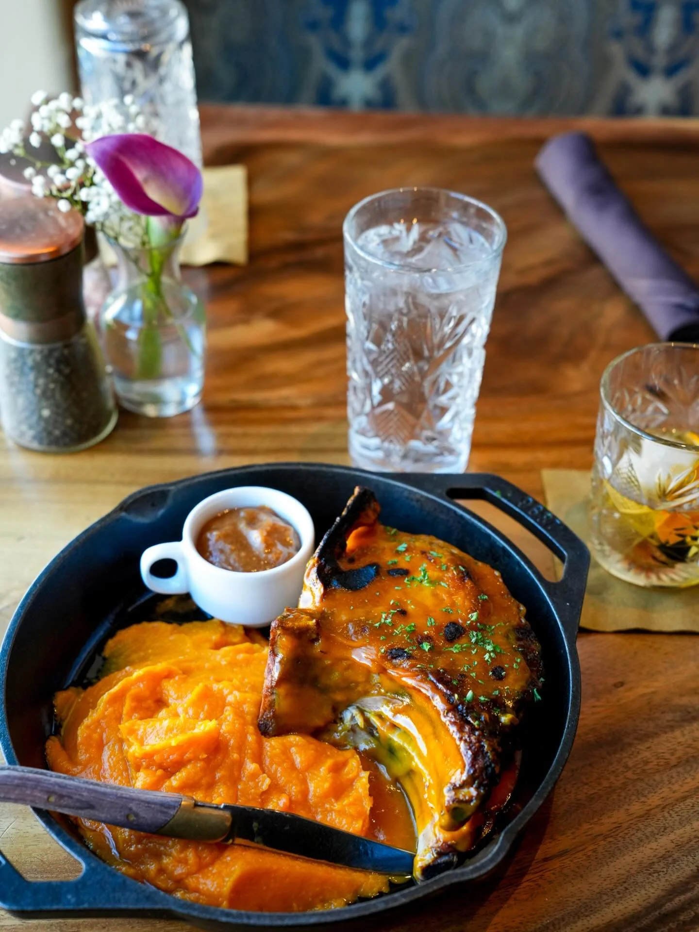A cast iron skillet with a cooked steak topped with sauce, served with mashed sweet potatoes. A small white ramekin of sauce is placed beside the steak. A fork is partially visible. The table has a glass of water, another drink, some flowers, salt and pepper shakers, a water pitcher, a rolled napkin, and a flower vase along with some decorative items.