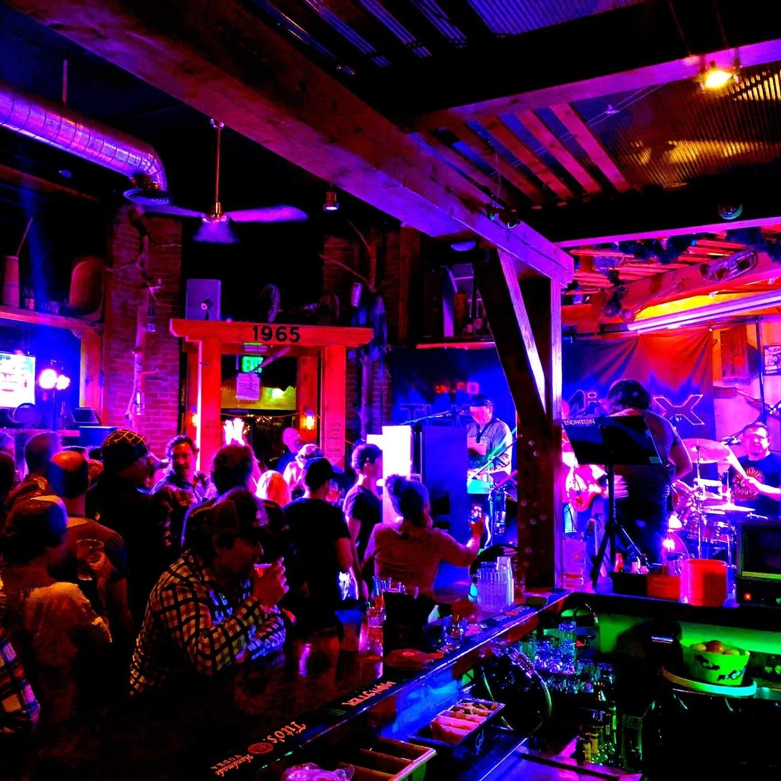 A lively indoor music venue with a band performing on stage and an audience enjoying the show. Neon signs, vibrant lighting, and a bar area are visible.