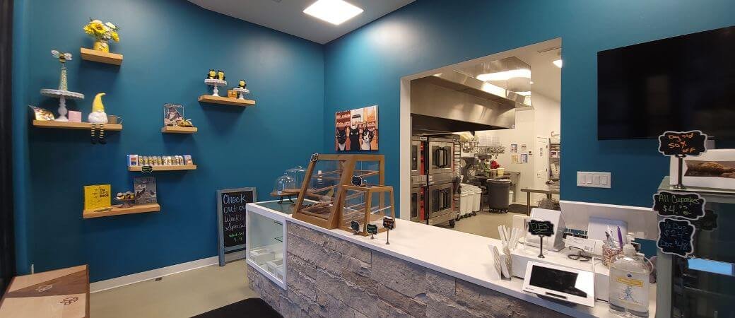 Interior of a small cafe or bakery with a blue wall, shelf displays with decorative items, a counter with a glass display case, and a kitchen visible in the background.