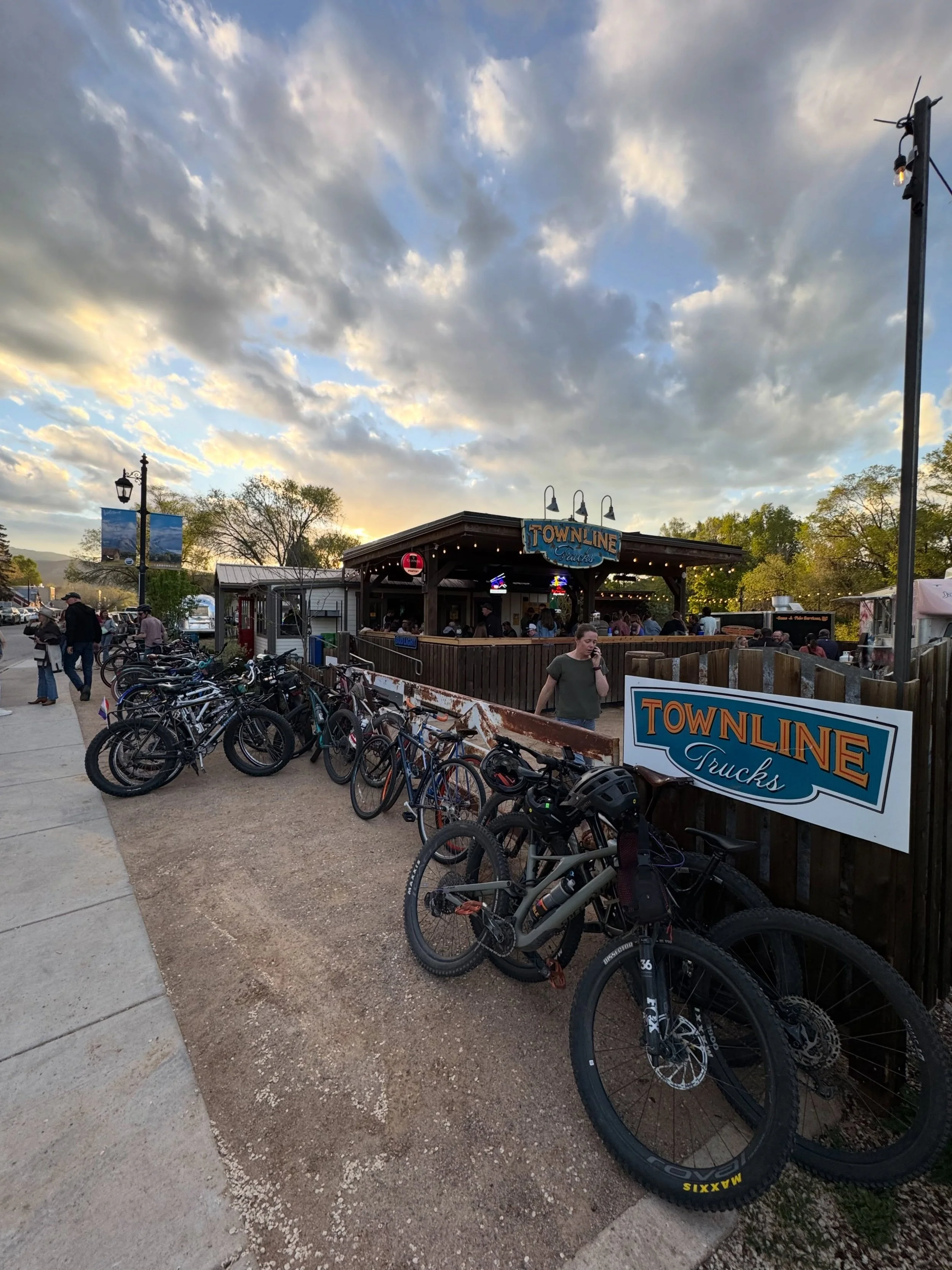 Outdoor scene at a restaurant called 'Townline Tucks' with a wooden structure and neon signs, a line of parked bicycles along a fence, some people walking and standing, trees in the background, cloudy sky, and street lamps.