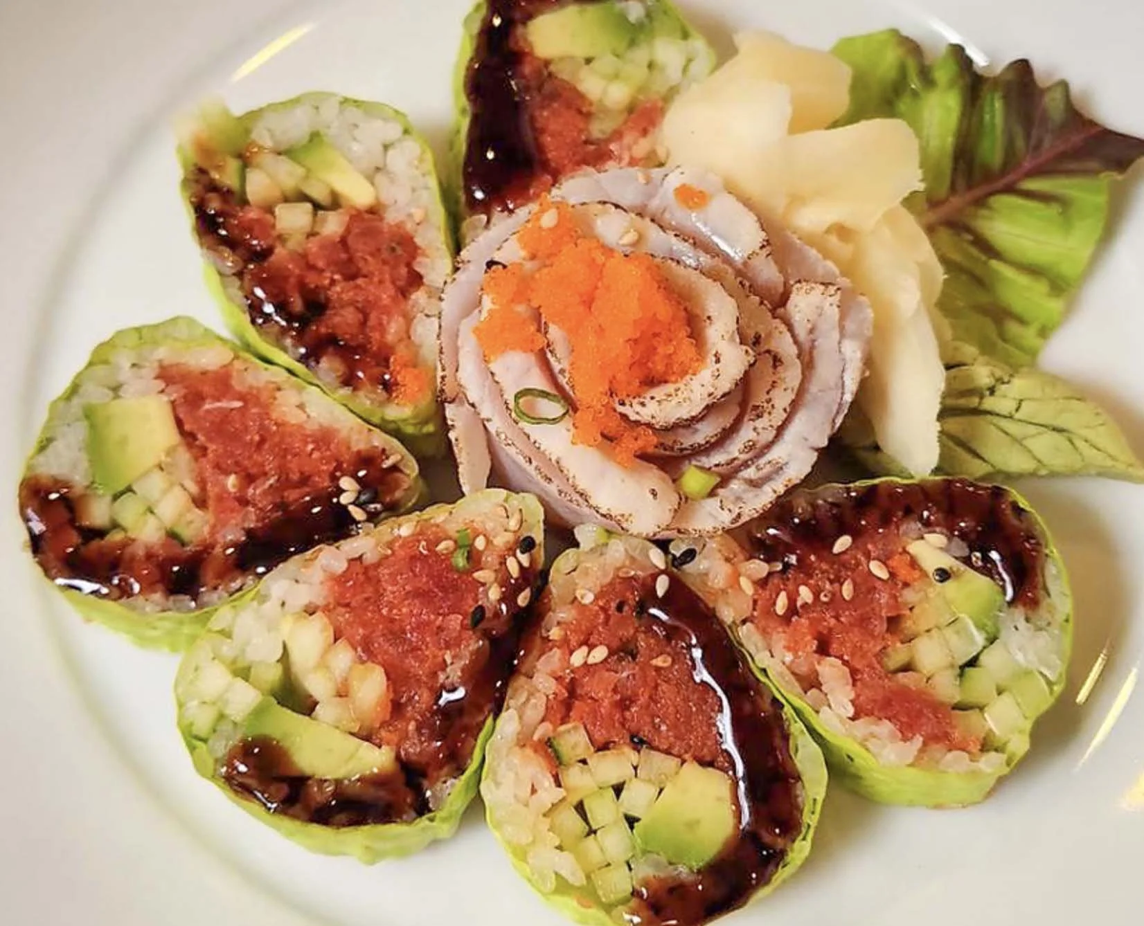 Close-up of sushi rolls filled with raw fish, chopped cucumber, topped with eel sauce and sprinkled with sesame seeds, garnished with a decorative carved vegetable flower and small slices of pickled ginger.