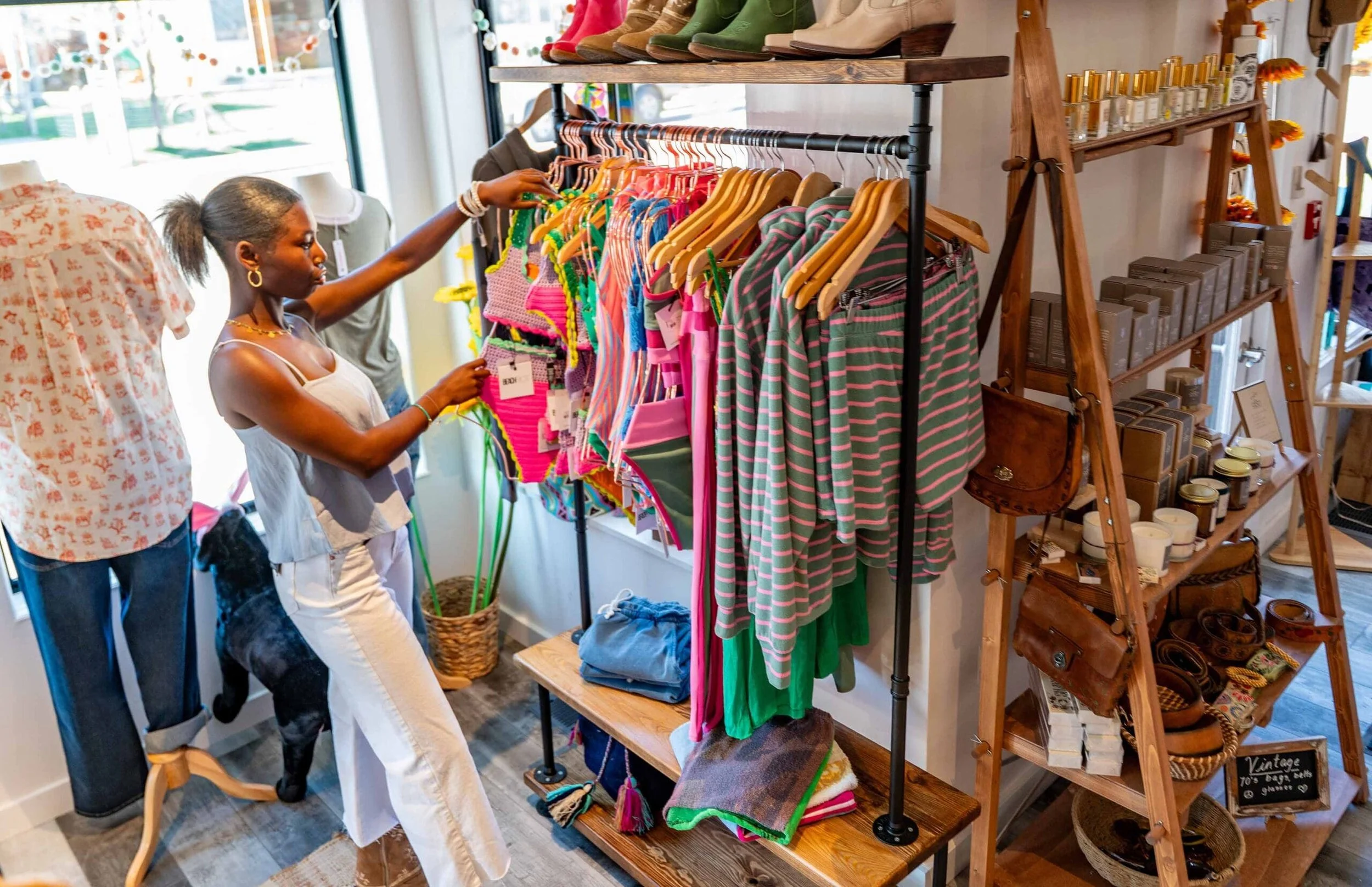 Two women shopping for clothes in a boutique, browsing colorful children's clothing on a rack near a window with sunlight. Shelves with perfume, candles, and vintage accessories are visible in the background.