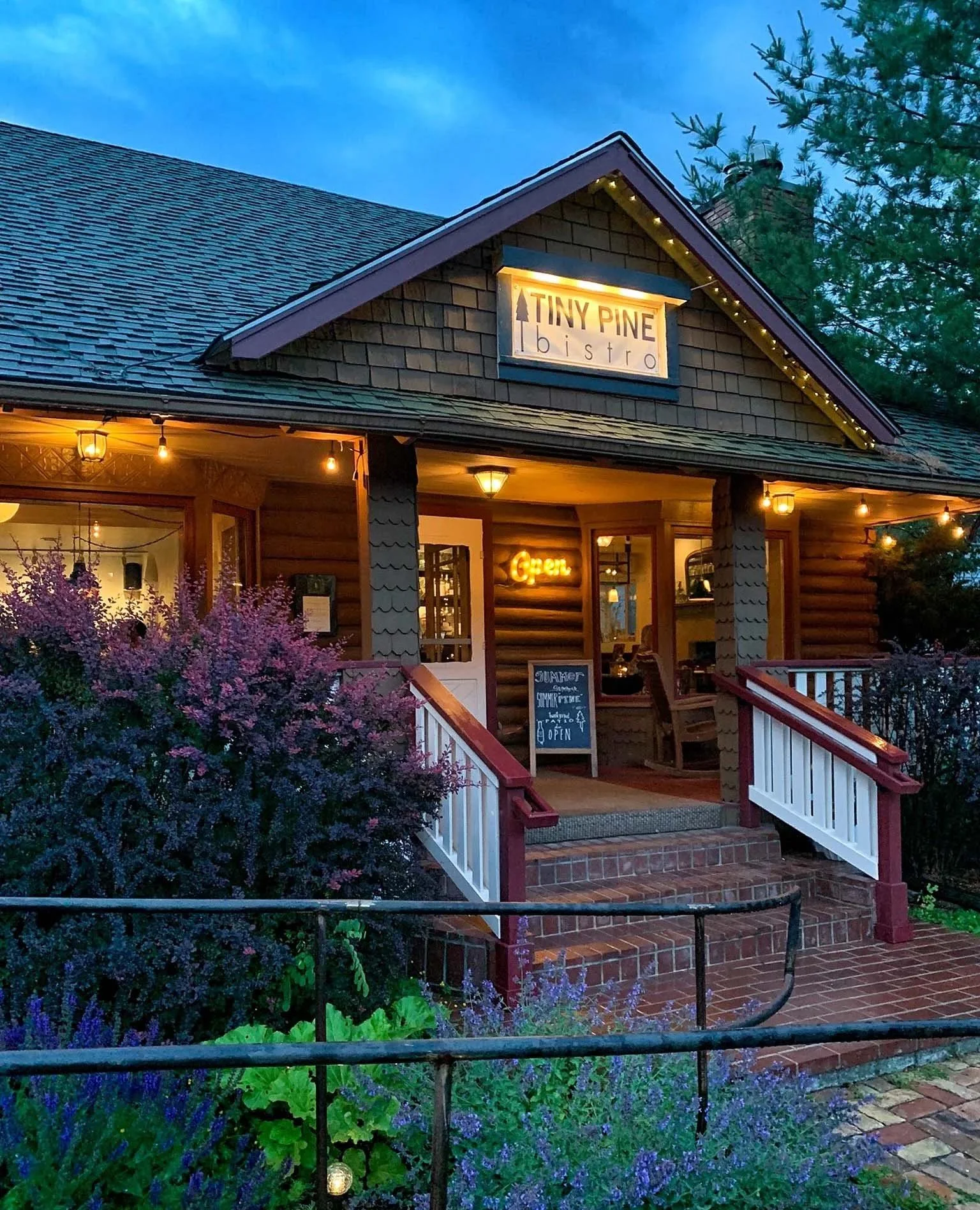 Exterior of a cozy wood cottage with a sign reading 'Tiny Pine Bistro' illuminated at dusk. The entry is decorated with string lights and there are flowering bushes in the foreground.