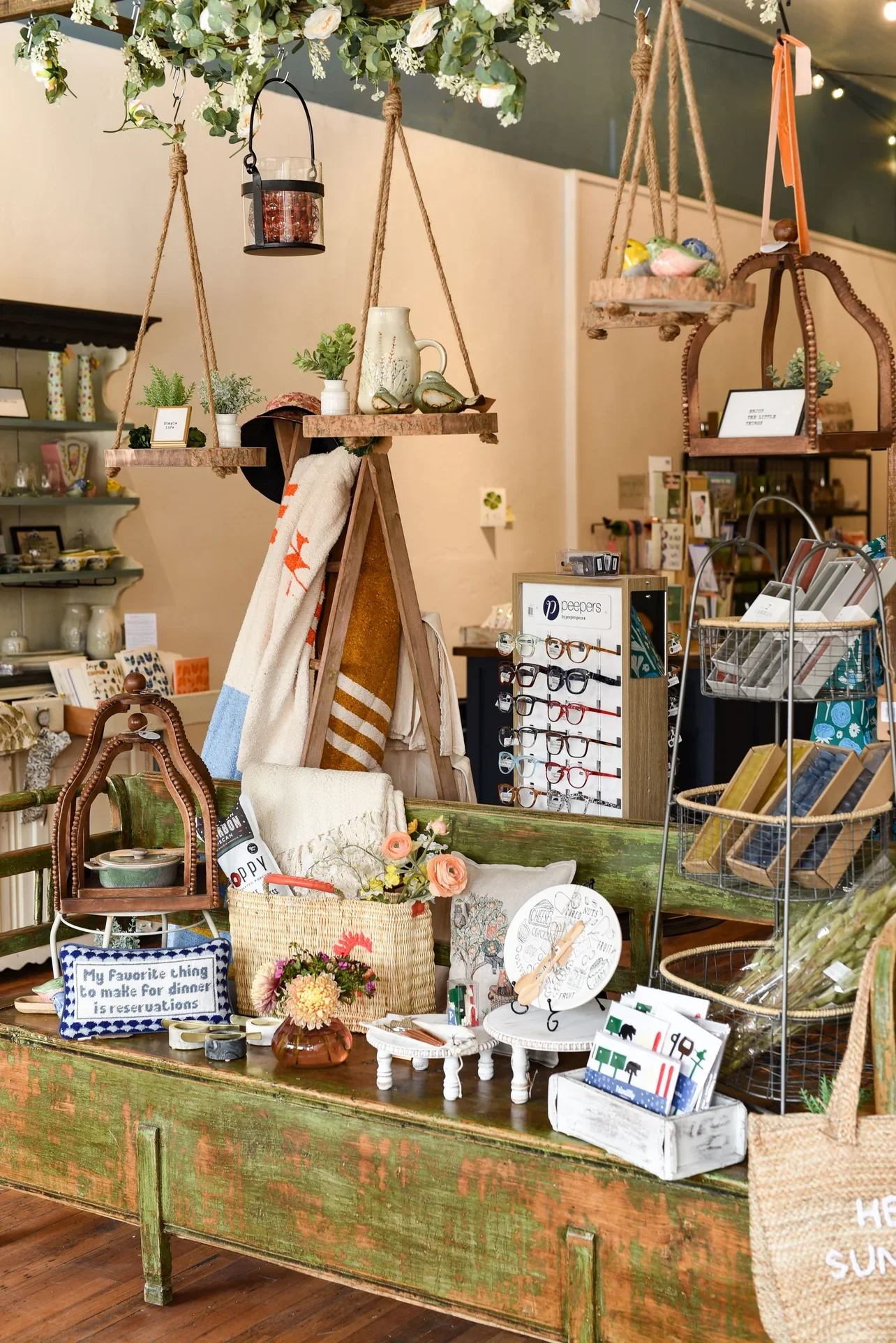 A display table with various home decor items, plants, and accessories at a market. Hanging shelves above hold decorative bird figurines, a pitcher, and small plants. There are eyeglasses, framed quotes, flowers, and small crafts on the table.