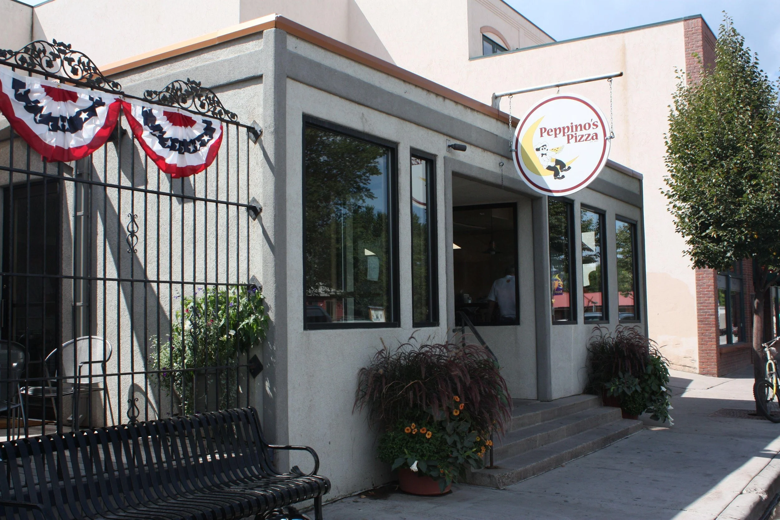 Exterior view of Peppino's Pizza restaurant with a hanging sign, decorative patriotic bunting on the fence, potted plants, a sidewalk, and entrance steps leading to the door.