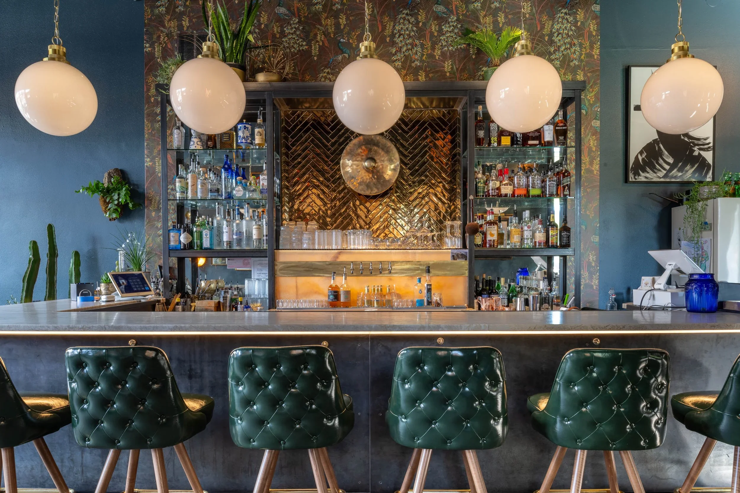 A bar with four large globe pendant lights hanging from the ceiling, a dark green wall on either side, a wall with botanical wallpaper behind the bar, and a framed black and white portrait to the right. The bar has various liquor bottles on shelves and glassware, and there are green leather bar stools in front with wooden legs.