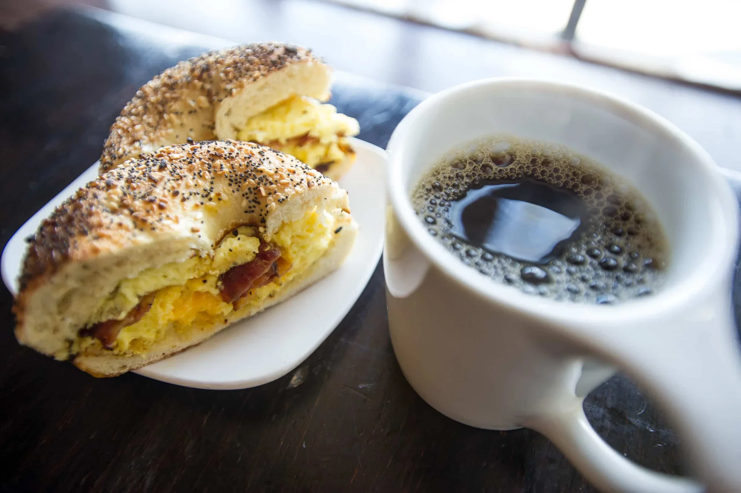 A breakfast consisting of a sausage and cheese breakfast bun with poppy seeds, a cup of black coffee, and a small serving of sliced potatoes in the background, on a dark surface.