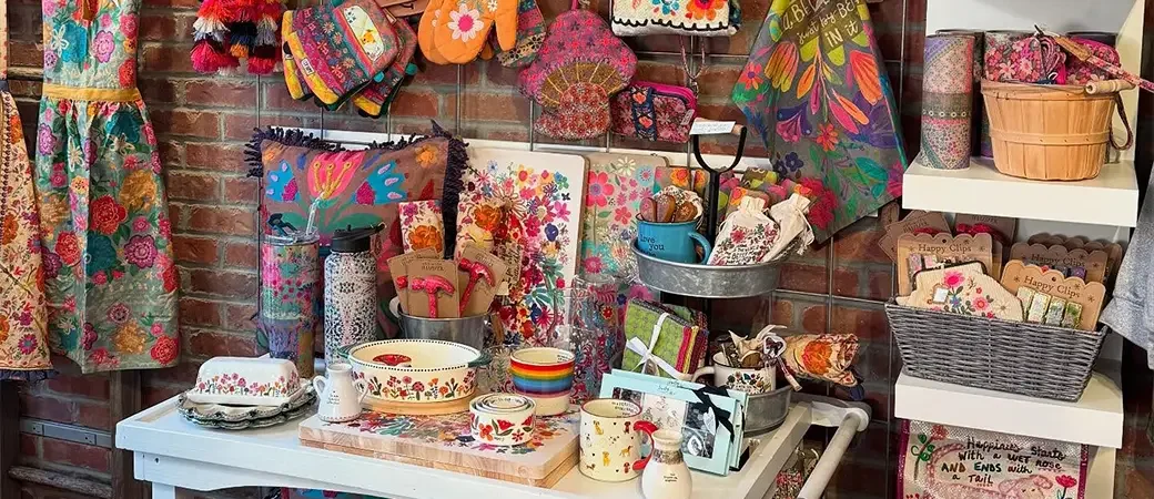 A display of colorful, floral-embroidered and patterned gifts, tableware, and accessories including mugs, plates, and decorative items on shelves and a table, with textile and bags hanging on a brick wall.