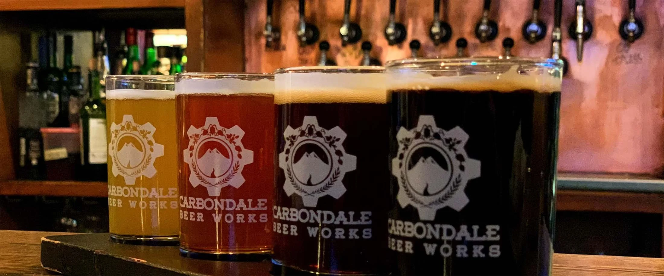 Four glasses of different types of beer on a wooden bar counter, with a beer tap and bottles in the background