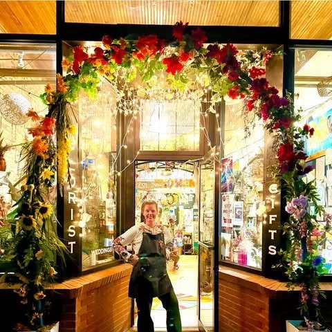 A woman standing under an archway decorated with colorful flowers and greenery at the entrance of a store or cafe, with bright sunlight behind.”}