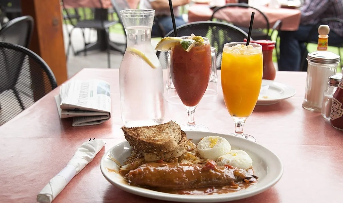 A breakfast plate with toast, eggs, and sausage gravy, along with three drinks in tall glasses and a pitcher of water on a pink tablecloth at an outdoor restaurant.