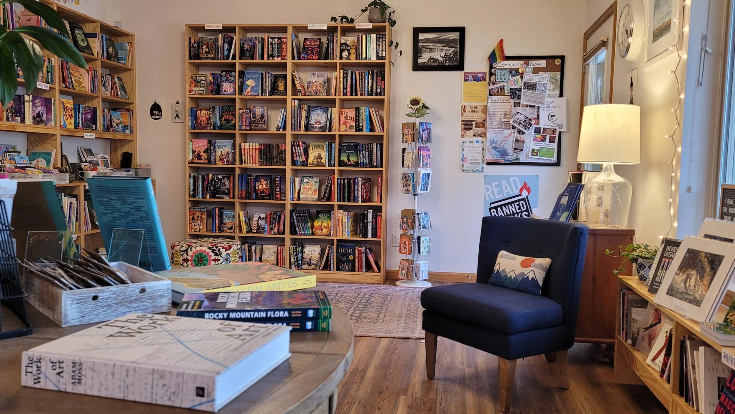 A cozy bookstore with wooden shelves filled with books, a reading nook with a black armchair, a lamp, and a bulletin board with flyers and a rainbow pride flag, decorated with string lights.