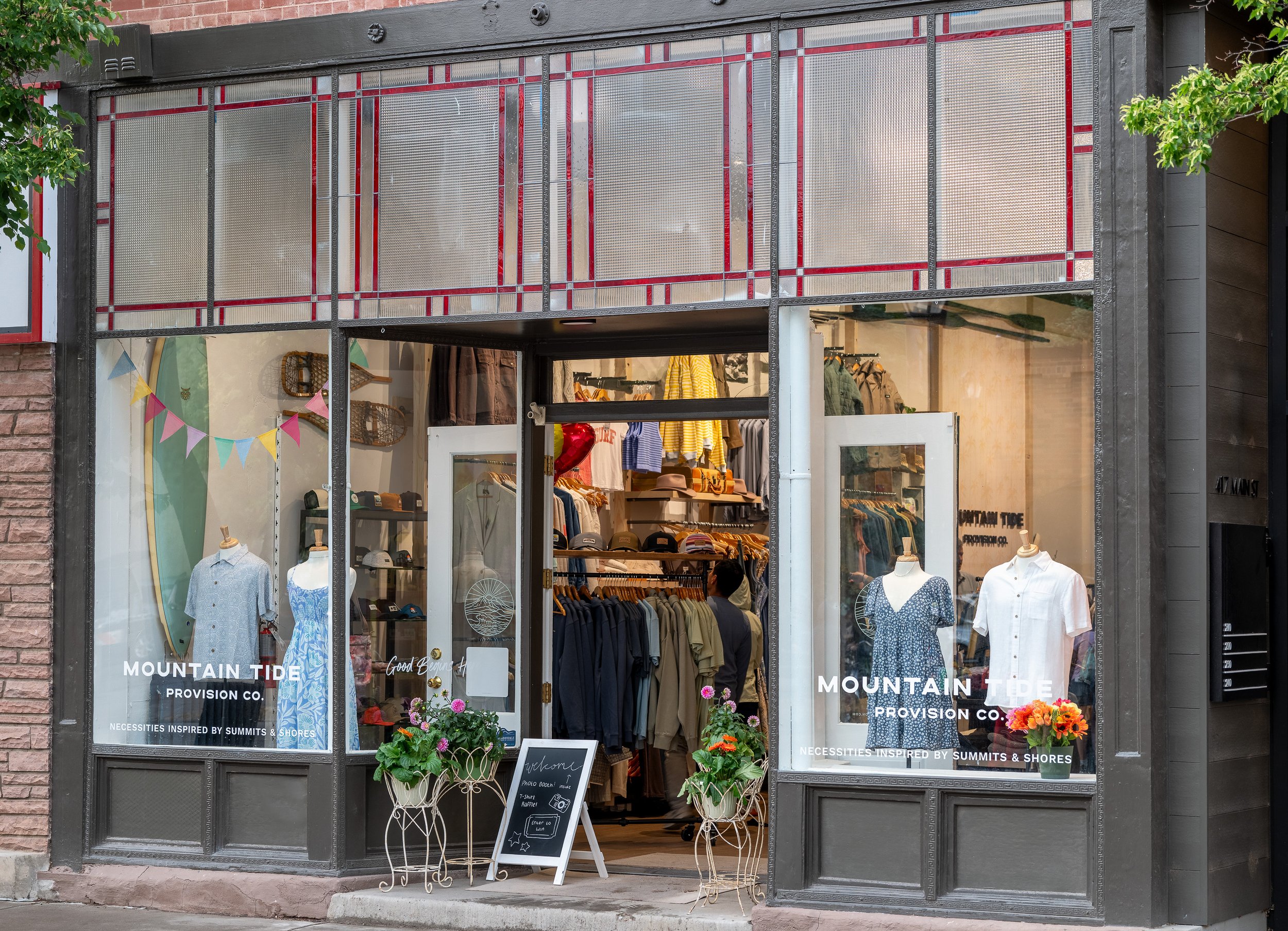 Storefront window display with clothing and accessories inside, including shirts, jackets, hats, and bags, with decorative plants and chalkboard signs outside. The store appears to be called 'Mountain Tide Provision Co.' with a slogan about necessities inspired by summits and shores.