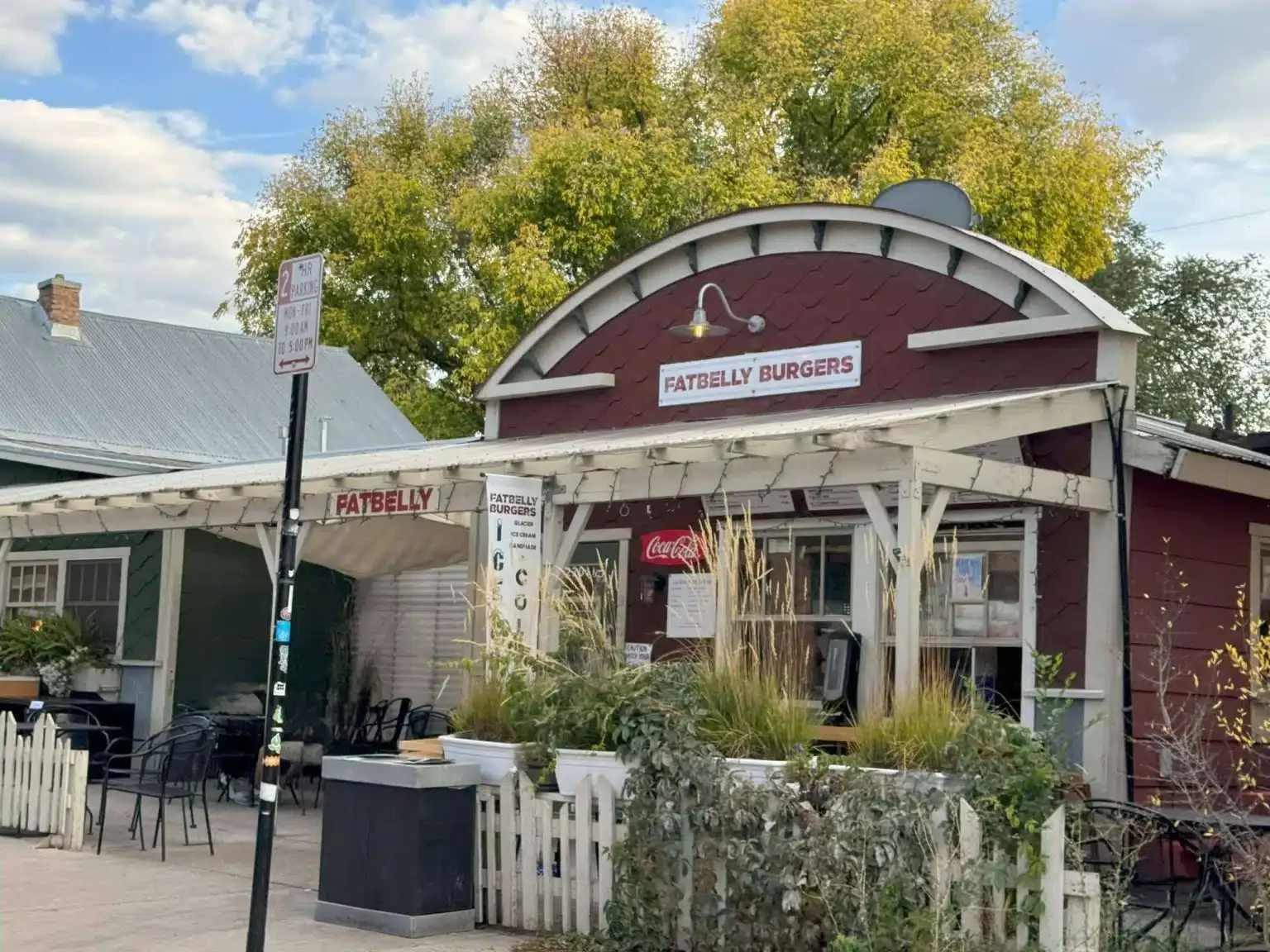 A small red restaurant named 'Fatbelly Burgers' with outdoor seating and a Coca-Cola sign. There are plants and a fencing in front, with a street parking sign and trees behind.