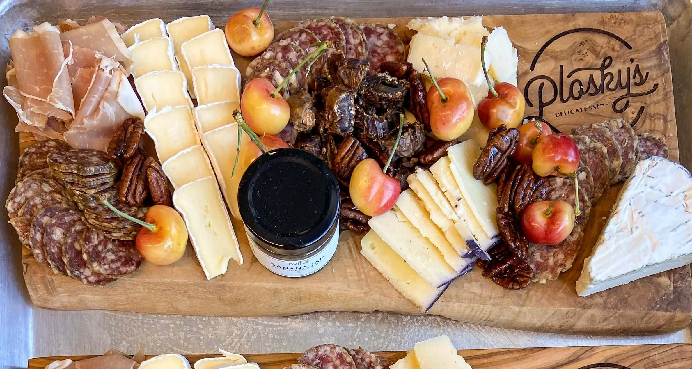 A wooden serving board with a variety of cheeses, cured meats, fresh cherries, pecans, and a jar of jam.