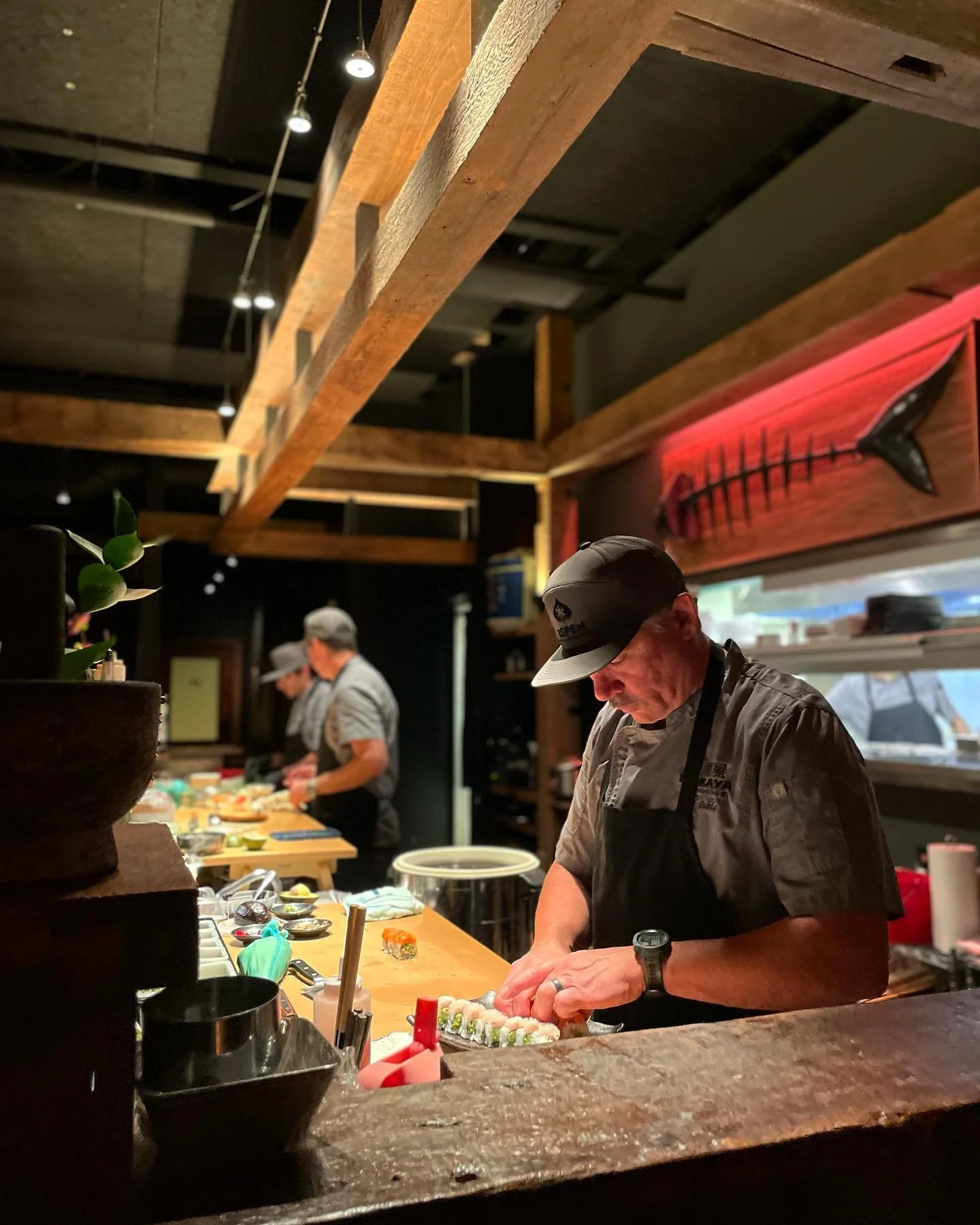 A chef preparing sushi in a restaurant kitchen, with other staff working in the background. The kitchen has wooden beams and a red whale wall art.