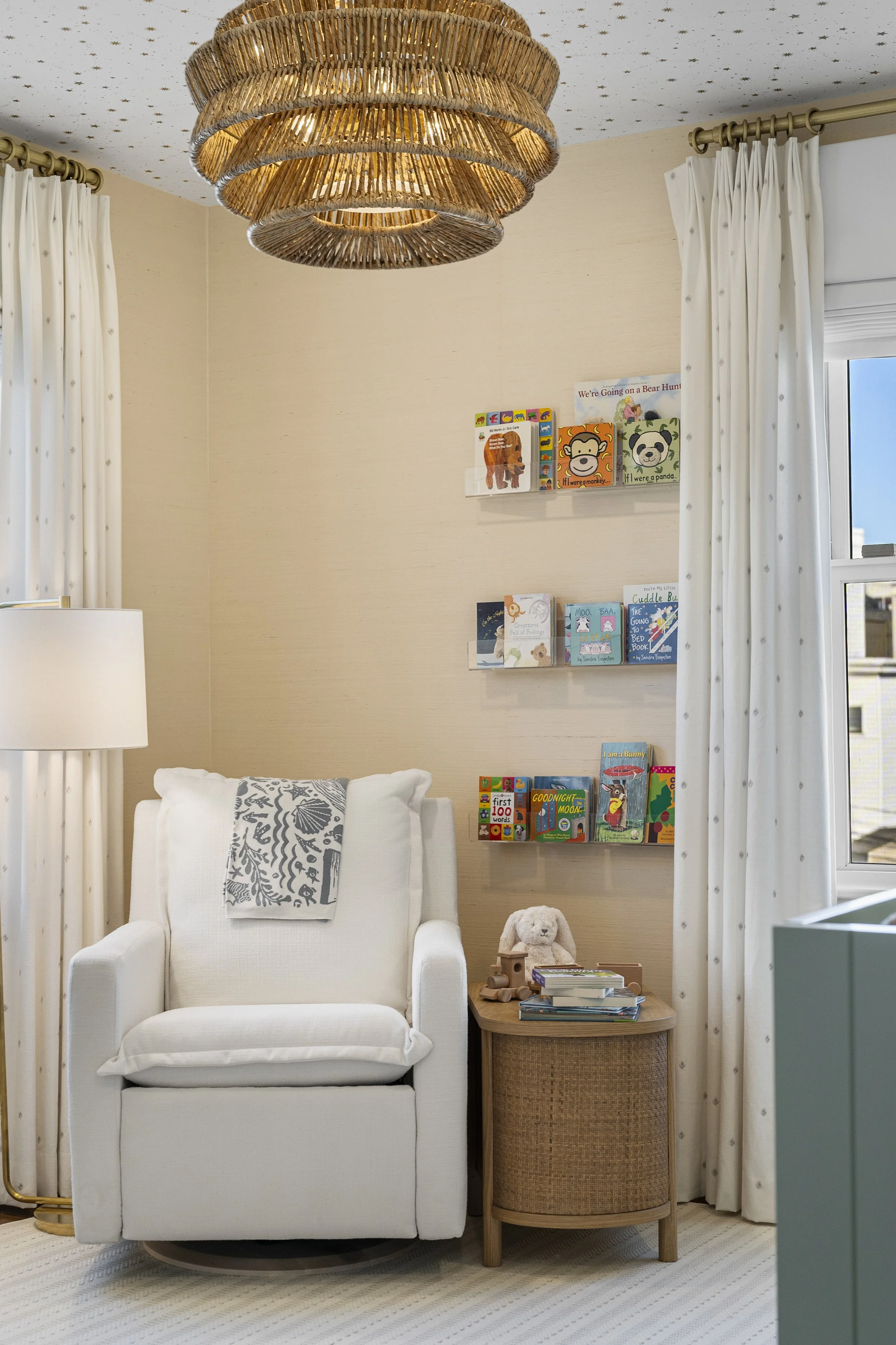 A cozy living room corner with a white armchair adorned with a gray patterned throw blanket. Next to the chair is a small wooden side table holding a teddy bear, books, and toys. Behind the chair, on the wall, are two shelves filled with children's b