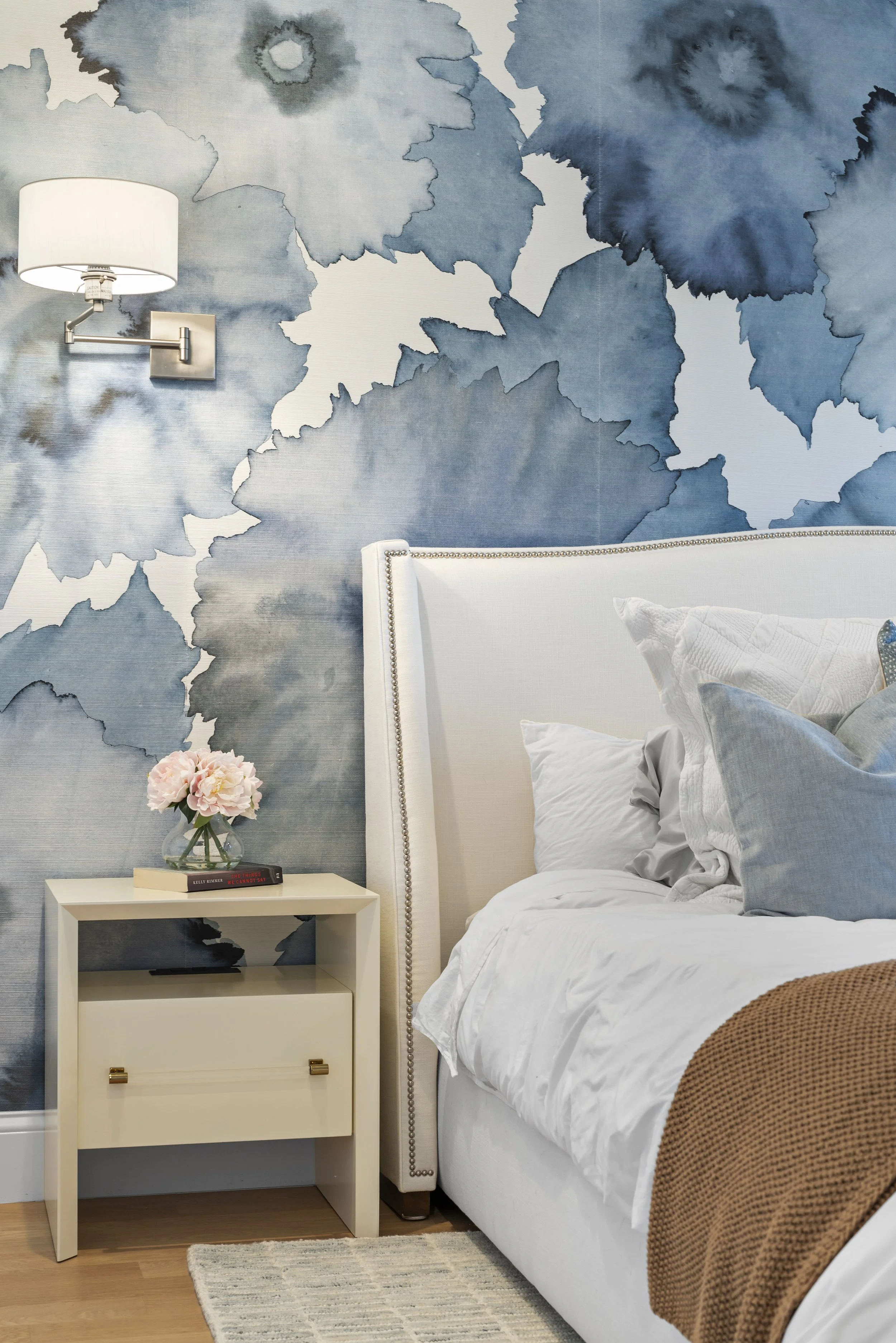 A bedroom with a white bed, a nightstand with a vase of pink flowers and books, a wall-mounted lamp, and blue watercolor floral wallpaper.