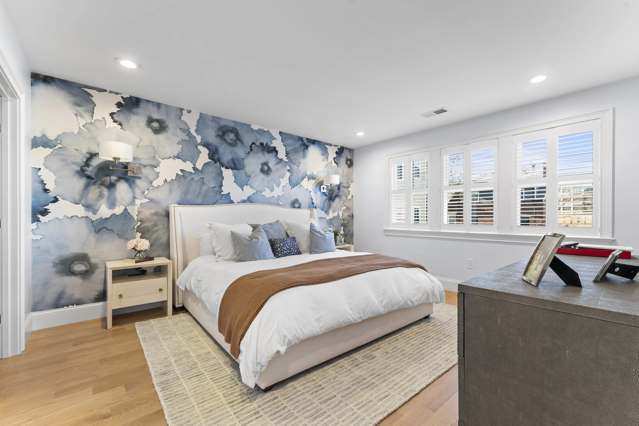 A bedroom with white walls, a window with shutters, and a bed with white linens and a brown throw blanket. There are two wall-mounted lamps, a nightstand with a flower arrangement, and a dresser with picture frames.