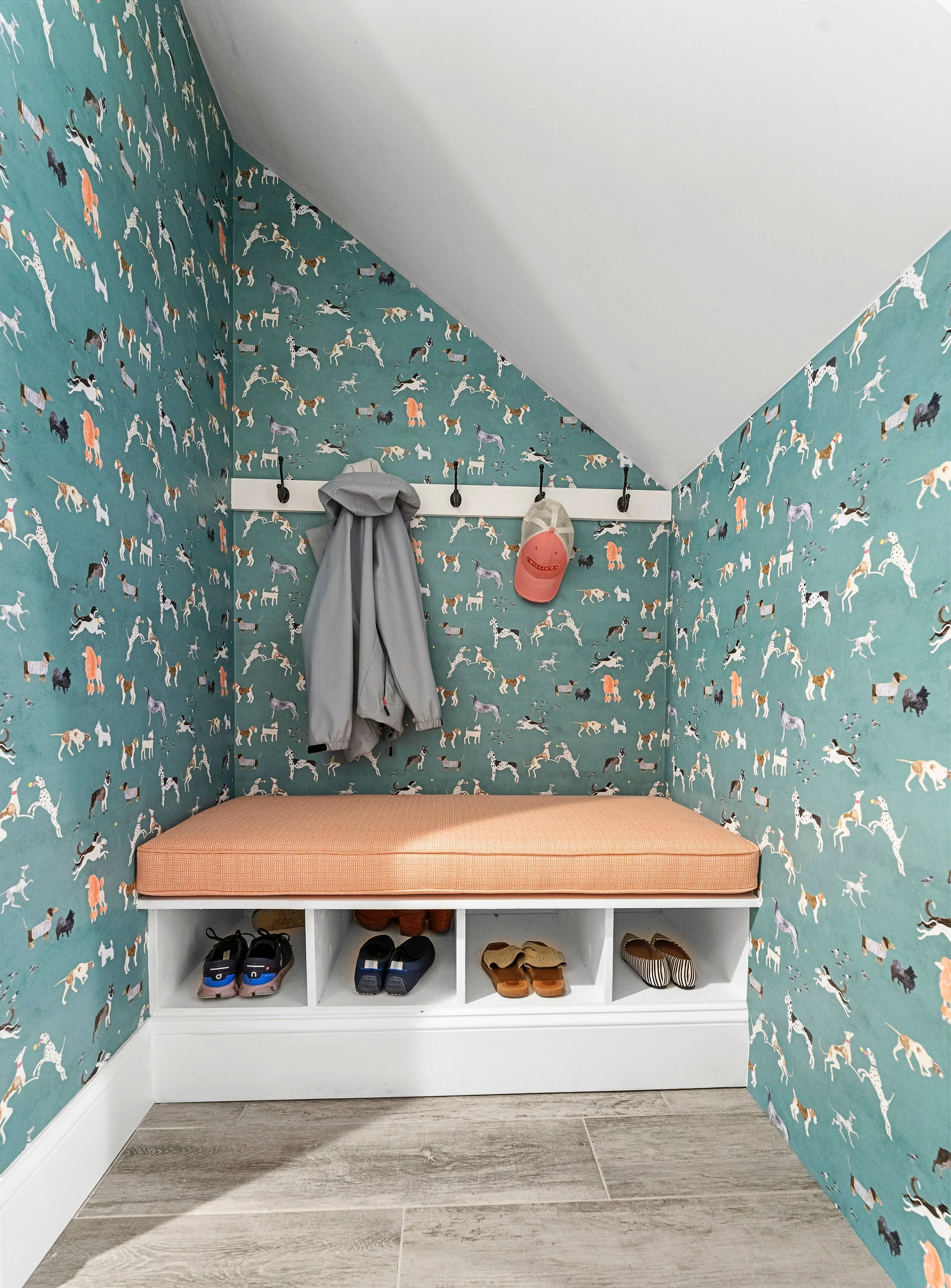 A small entryway nook with a wall of dog-patterned wallpaper, a white shoe storage bench with an orange cushion, and hooks holding a gray jacket and a pink cap. Shoes and sneakers are stored in the cubbies beneath.