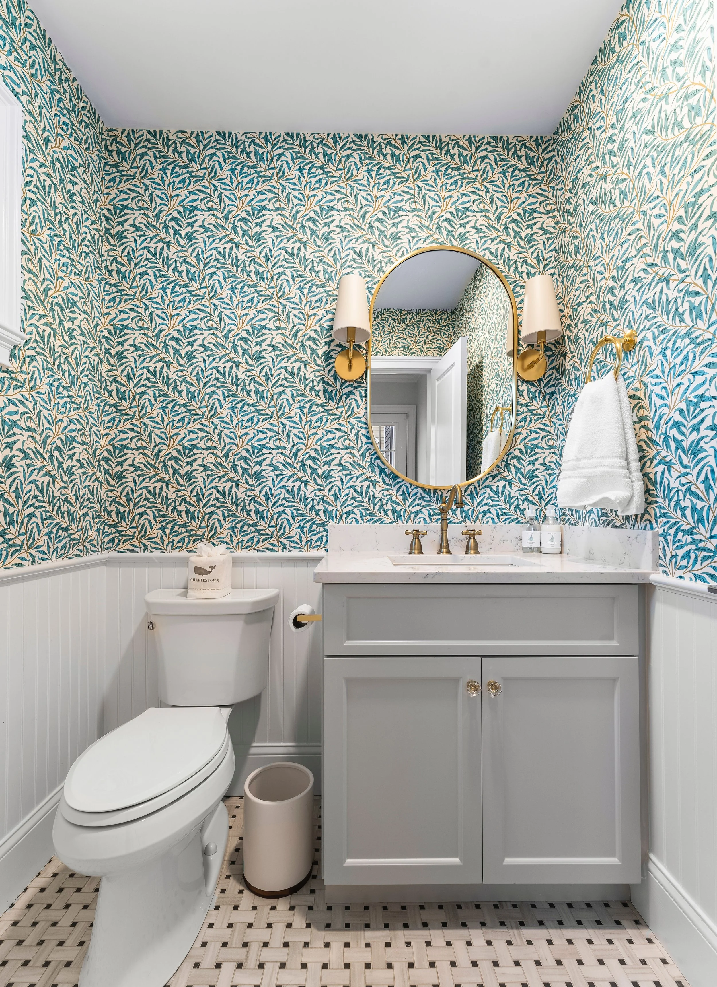 Bathroom with wallpaper featuring a leafy pattern, a white vanity cabinet with a marble countertop, a round mirror with a gold frame, two wall-mounted sconces with white shades, a white towel hanging on a gold hook, a toilet with a tissue box on top,