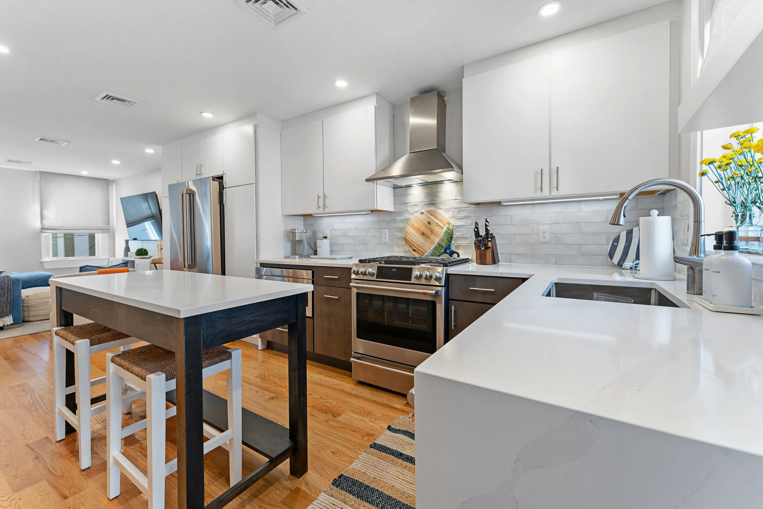 Modern kitchen with white cabinets, stainless steel appliances, a wooden cutting board, black knife set, and white marble countertops. There is a window with yellow flowers, a coffee or soap dispenser, and a view of a living room with a blue seating 