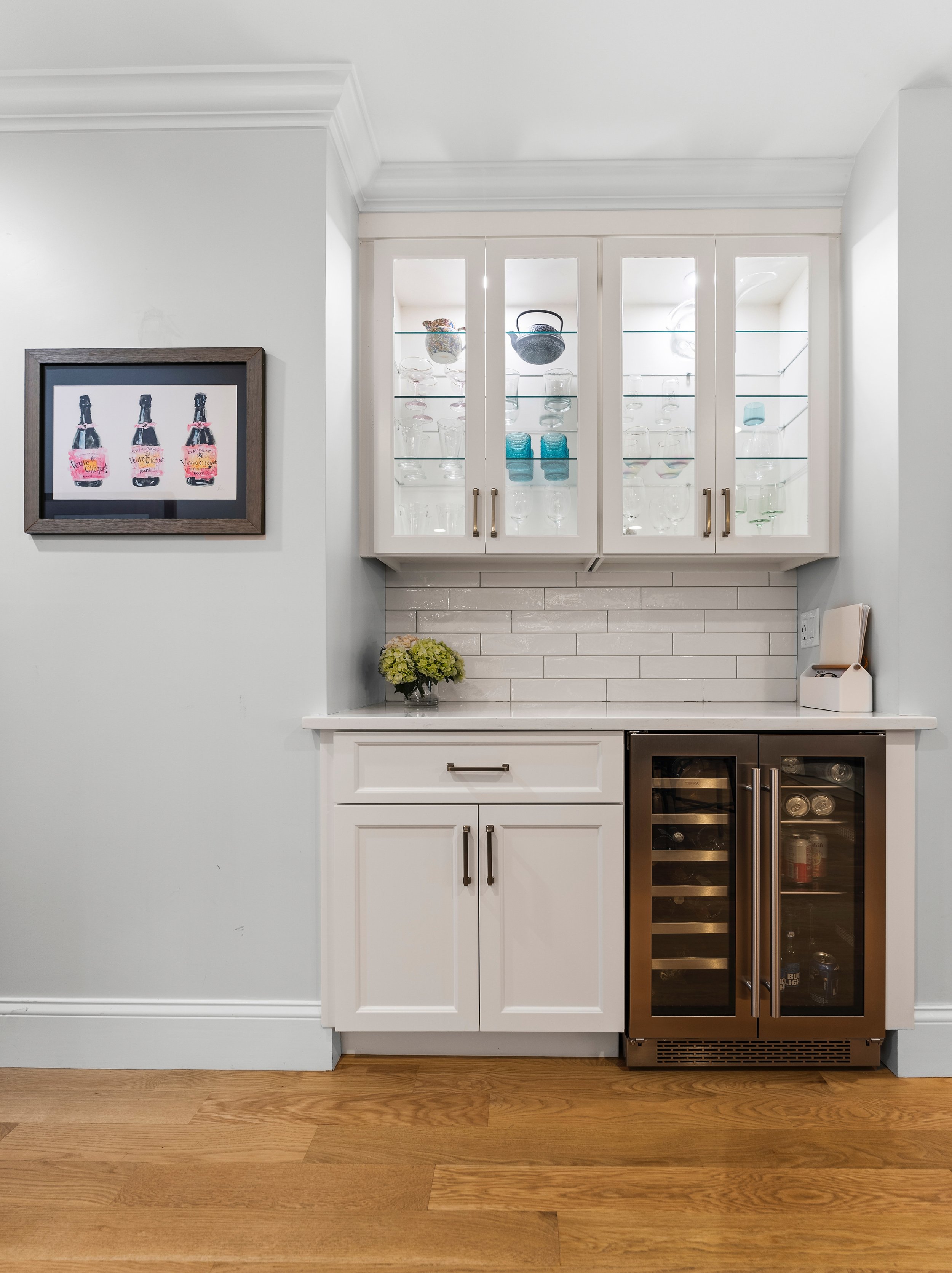 White kitchen cabinet with glass doors displaying glassware, a small wine fridge to the right, a framed picture of three bottles on the wall to the left, a small floral arrangement on the counter, and a plug-in paper towel holder on the right side.