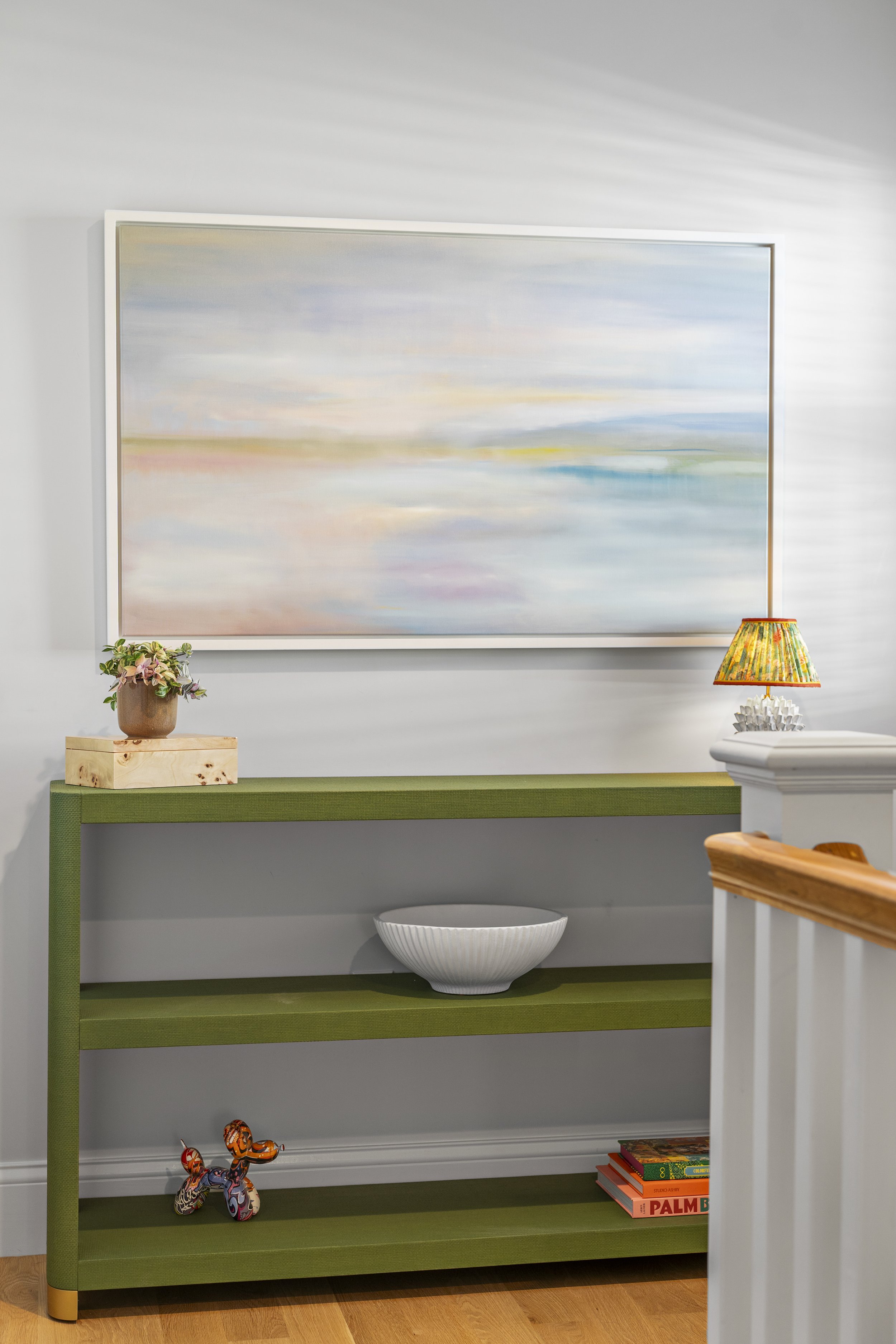 A green and white bookshelf with two shelves, decorated with toys, books, and decorative items, placed in a room with a large abstract painting on the wall and a lamp on a side table.