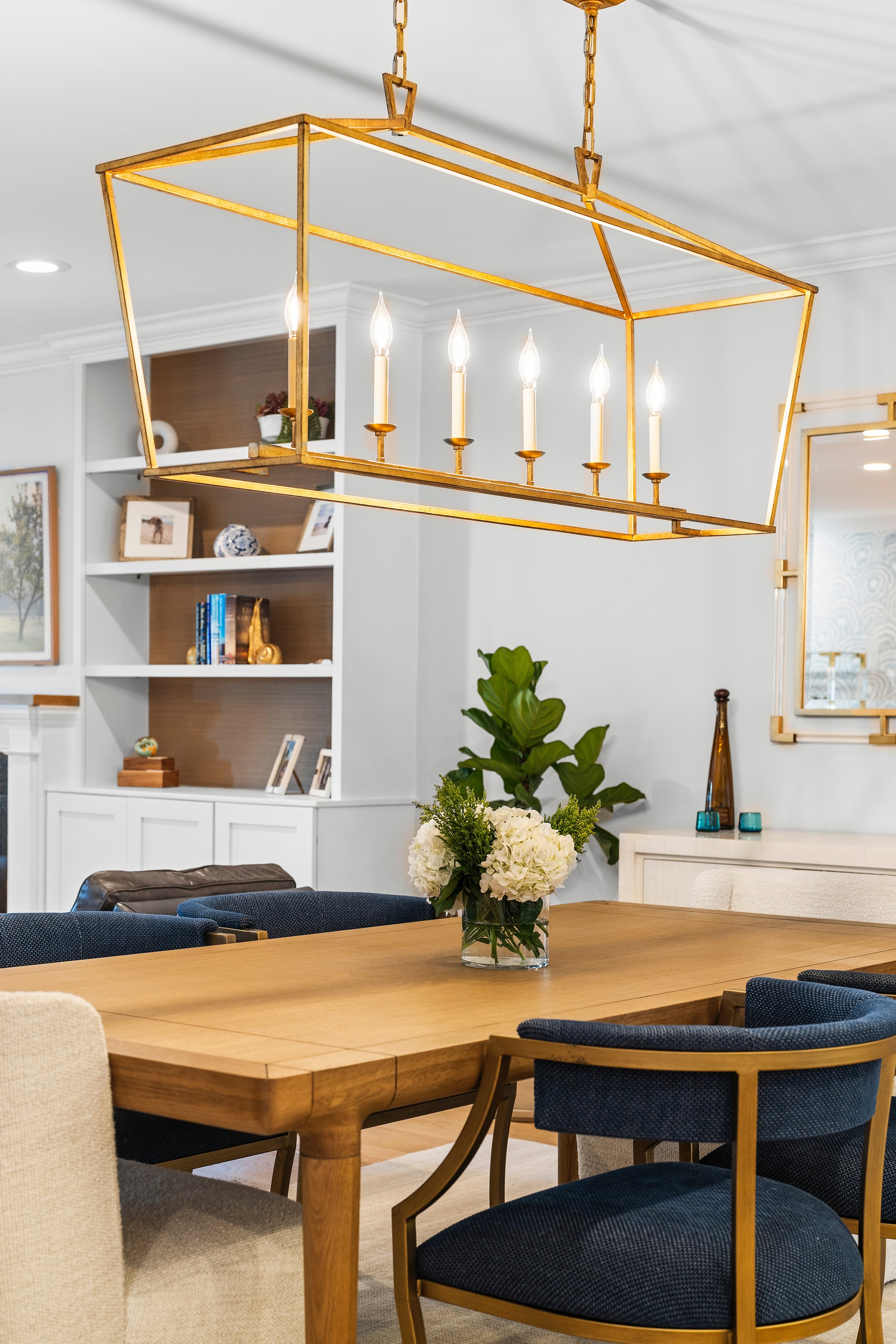 A modern dining room with a wooden table and blue upholstered chairs, decorated with a vase of white flowers, and a brass chandelier with candle-style lights hanging above.