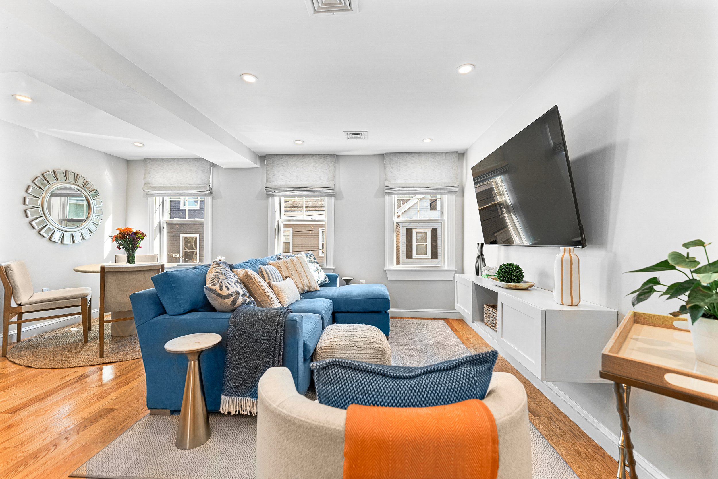 Living room with a blue sofa, beige chairs, a round wooden side table, and a beige pouf. There are three windows with shades, and a wall-mounted TV. Decor includes a round mirror, colorful flowers, and vases.