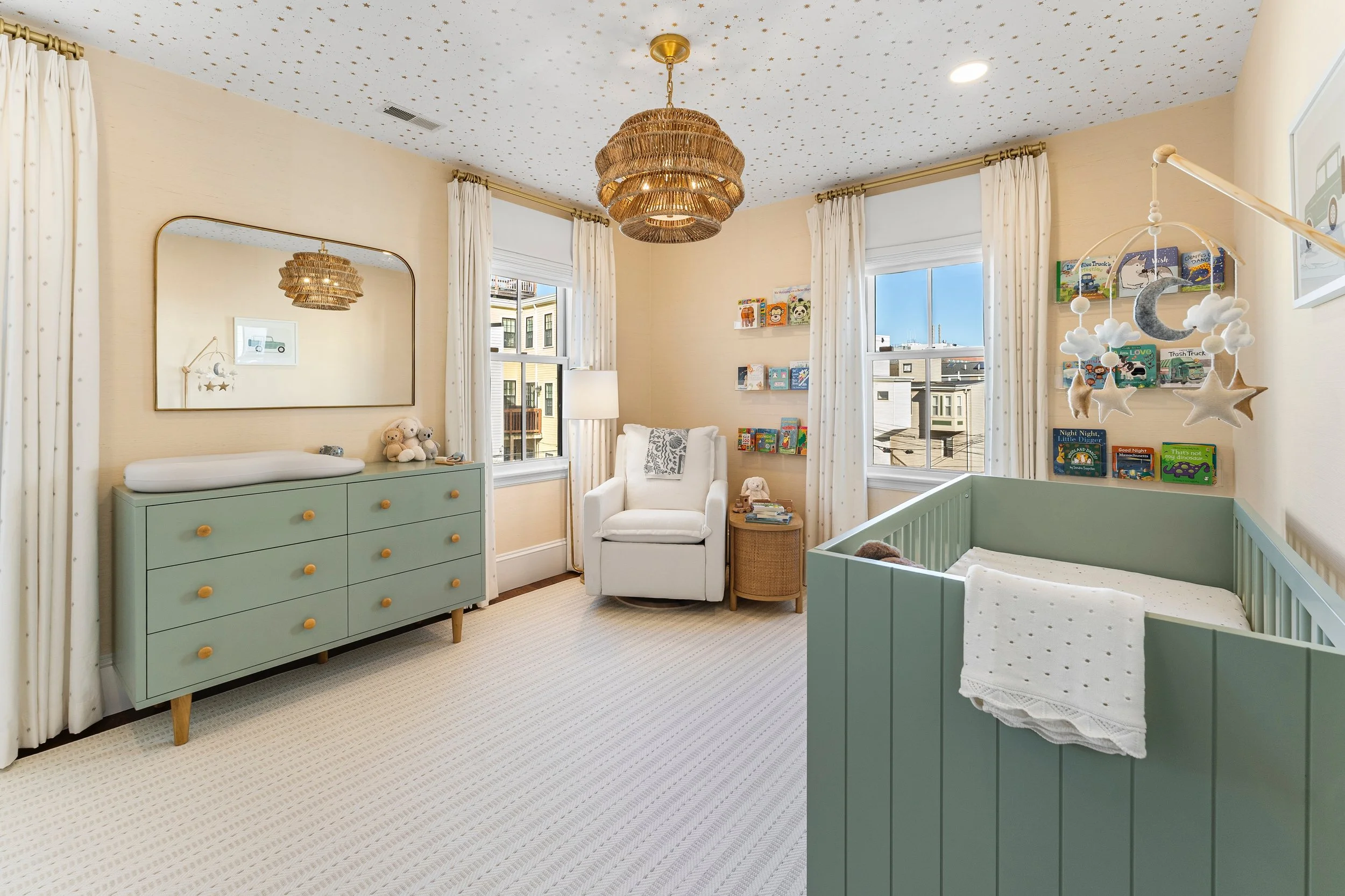 A nursery room with a green crib, a white armchair, a green dresser with stuffed animals on top, a hanging mobile with stars and clouds, books on wall shelves, a large mirror, a window with white curtains, ceiling with stars, and wicker pendant light