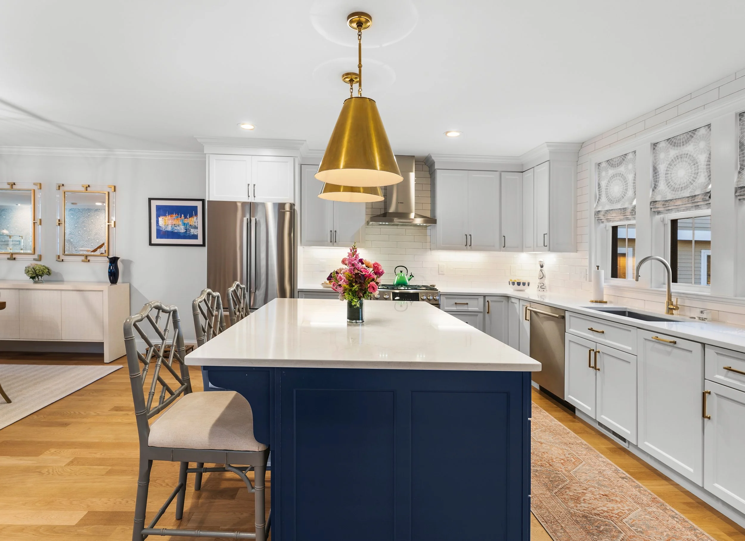 Modern kitchen with white cabinets, stainless steel appliances, and a blue island. Brass pendant lights hang above a white countertop with a pink flower bouquet, and framed artwork on the wall.
