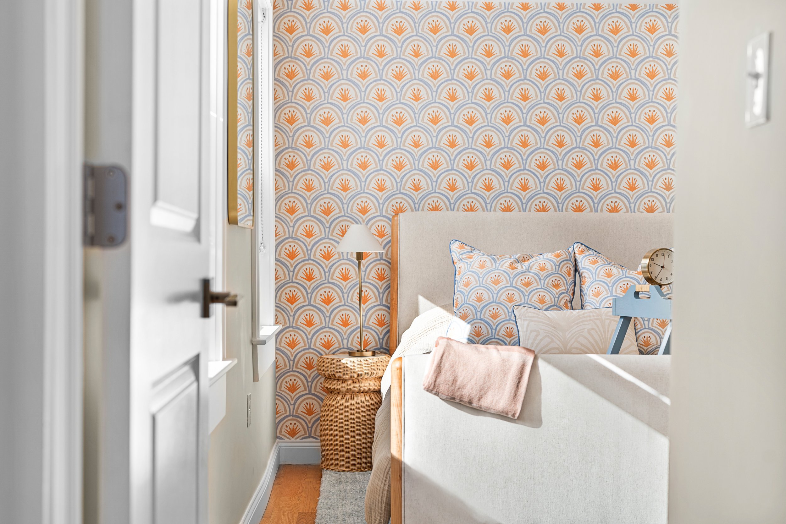 A bedroom decorated with retro-style wallpaper featuring orange and blue fan motifs, a beige upholstered bed with patterned pillows, a wicker basket, a white bedside lamp, and a small blue high chair. The room has natural light coming through a windo