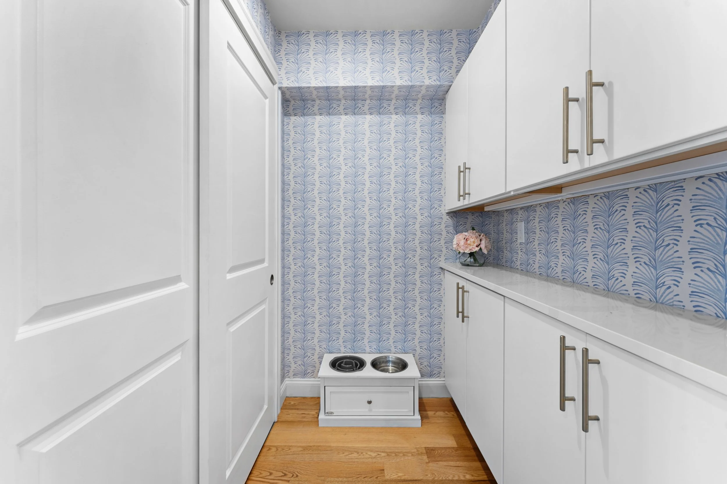 Small kitchen corner with white cabinets, a blue patterned wallpaper, a small white cabinet with a flower vase, and a two-burner hotplate.