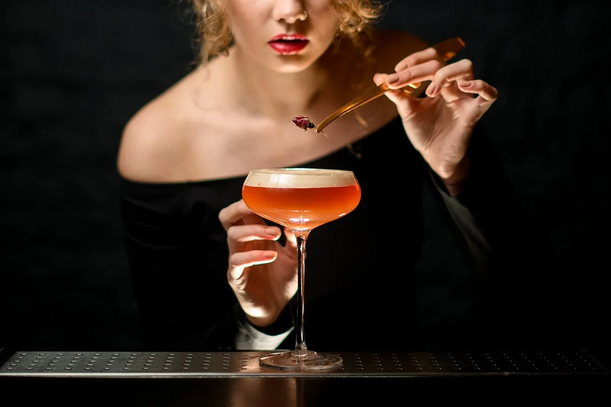 A woman with curly hair, red lipstick, and a black off-the-shoulder top holds a cocktail glass with amber-colored drink and foam topping, using a spoon to add a cherry garnish.
