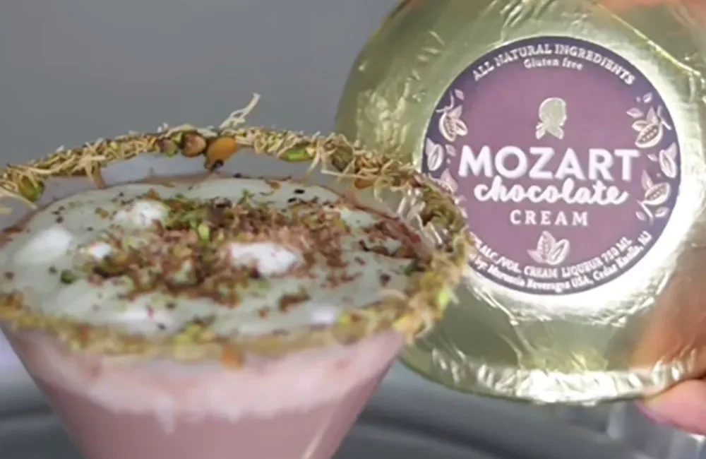 A glass of pink milkshake topped with whipped cream and chocolate shavings, with a package of Mozart chocolate cream in the background.