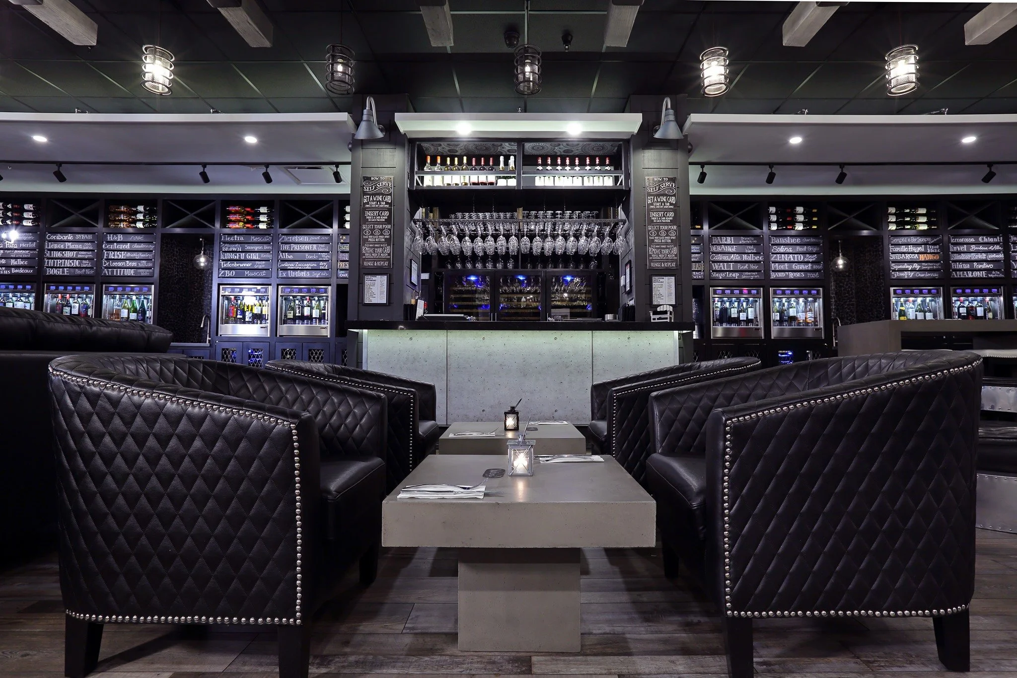 Interior of a modern bar or lounge area with black quilted leather chairs, a concrete table with a candle, and a bar in the background displaying wine bottles and glasses.