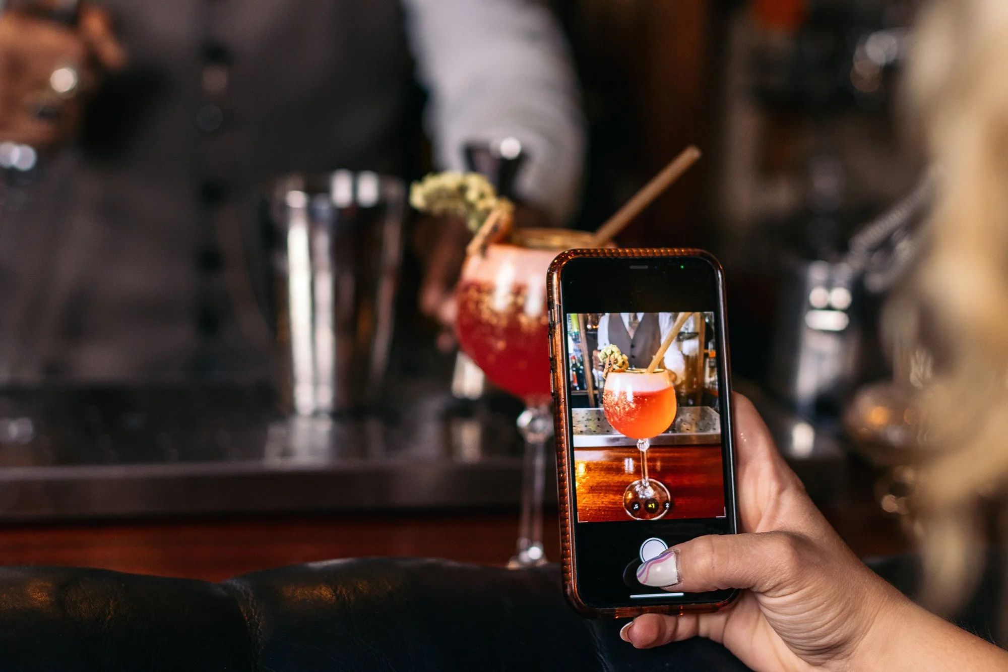 A person holding a smartphone taking a photo of a cocktail in a bar or restaurant. The cocktail is in a coupe glass and garnished with a lemon peel and cinnamon stick. The background is blurry with some drink shakers visible.