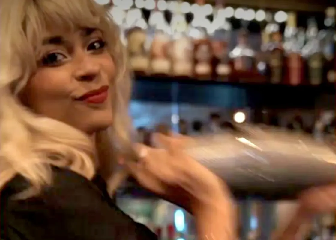 A woman with blonde hair and red lipstick behind a bar, holding a tray of drinks.
