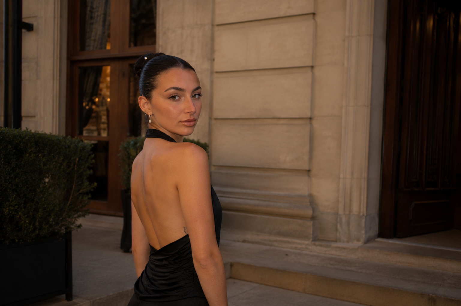 A woman in a black evening gown with an open back, standing outside a building with stone walls and steps, looking over her shoulder.