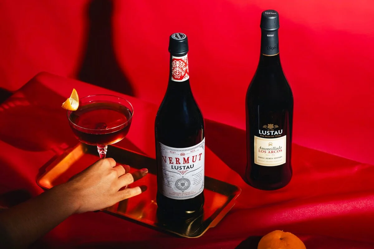 A hand holding a glass of red wine with a lemon wedge garnish, next to two bottles of wine on a red satin cloth background.