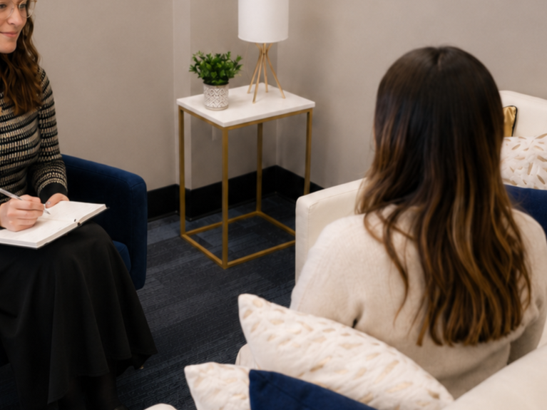 Two women sitting in a therapy or counseling session in a cozy, modern room. One woman is taking notes, while the other has her back to the camera. There is a white nightstand with a plant and a lamp between them.